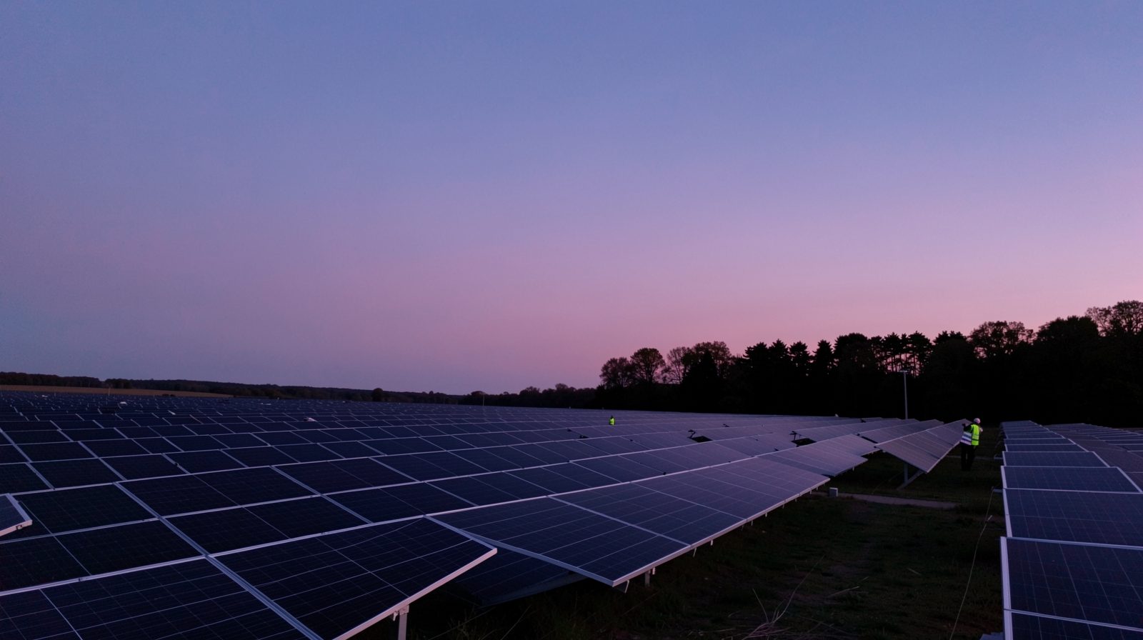 Rows of dark solar panels at blue hour, with a technician in a high-visibility jacket walking between them